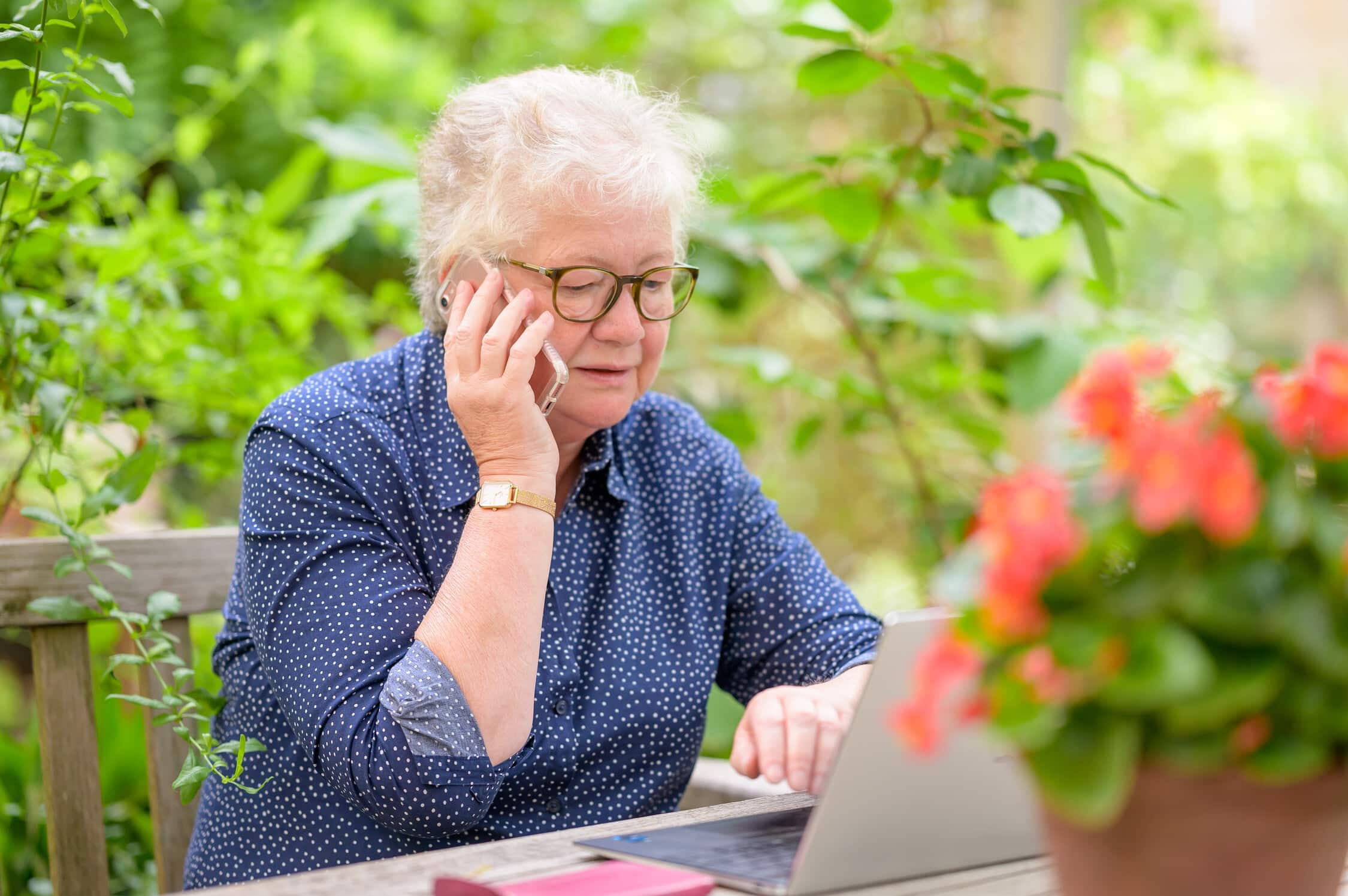 Elderly person on phone, using laptop at garden table.