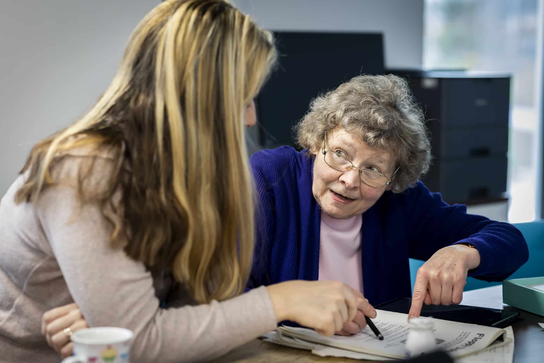 Two women sit at a table, reviewing documents together, one pointing and the other holding a pen.
