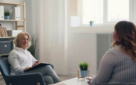 Two women are seated in an office, engaged in conversation. One is taking notes.