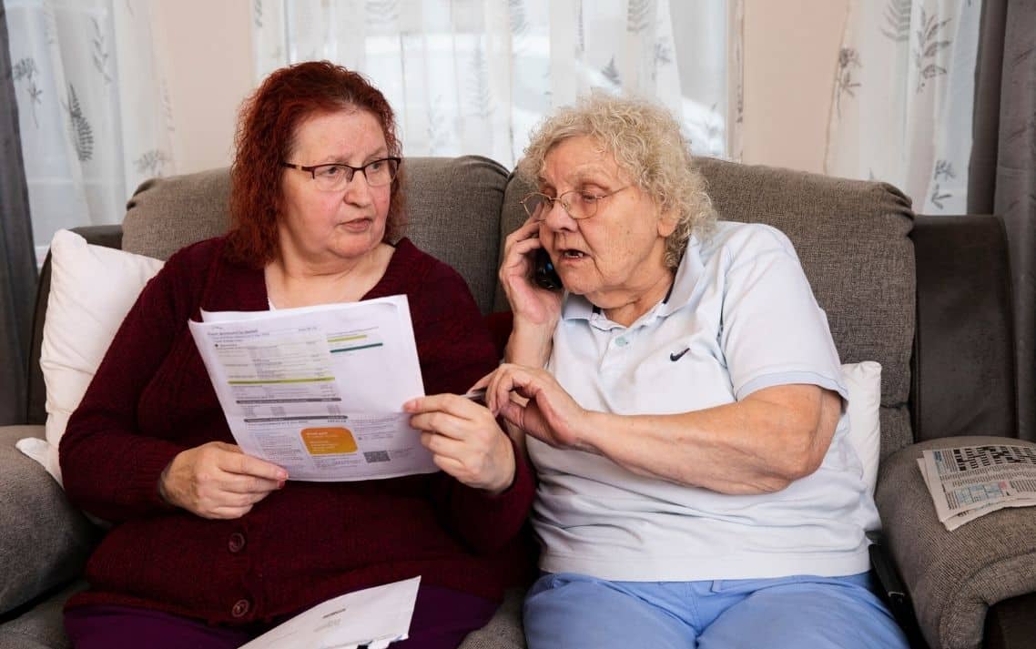 Two elderly women sit on a couch, one holding a bill, the other speaking on a phone.
