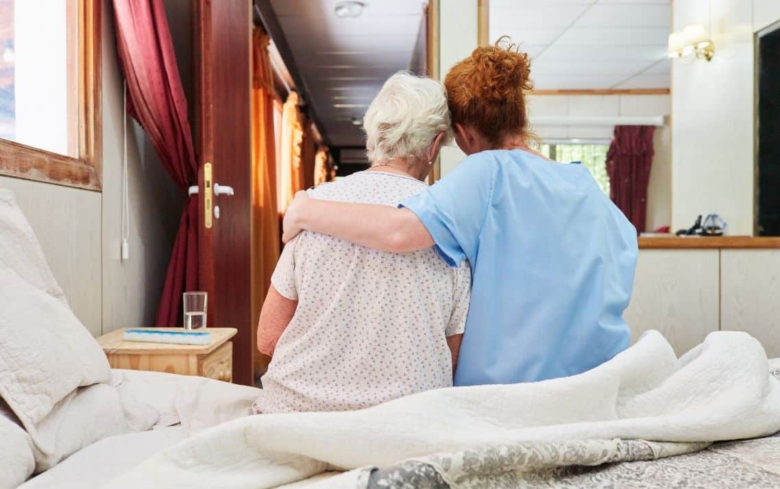 Caregiver and elderly woman sitting together, embracing on a bed.