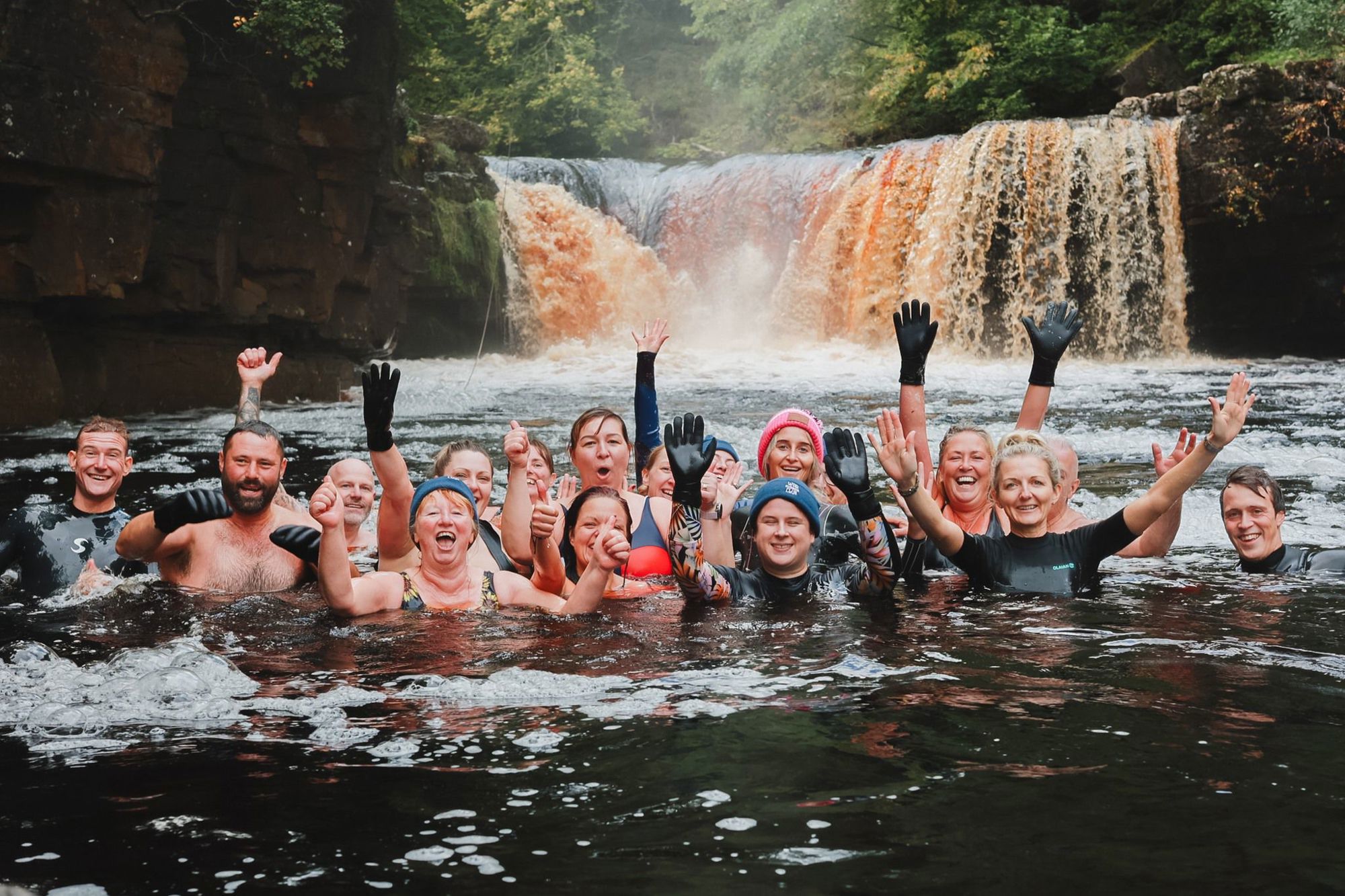 Group of people smiling and waving in a river near a waterfall.
