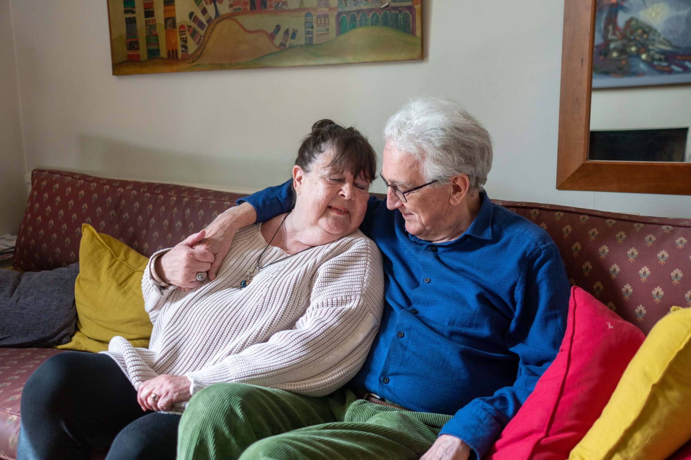 Elderly couple sitting on a sofa, embracing and smiling warmly at each other.