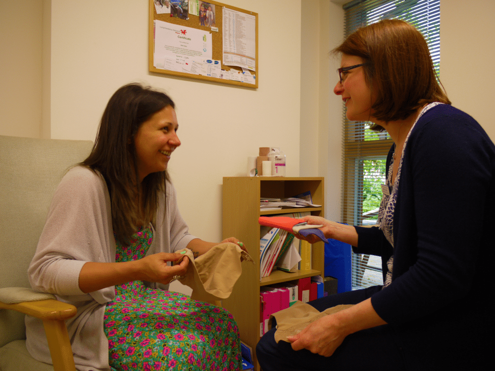 Two women sitting and smiling while holding fabric swatches in a well-lit room with shelves and a bulletin board.