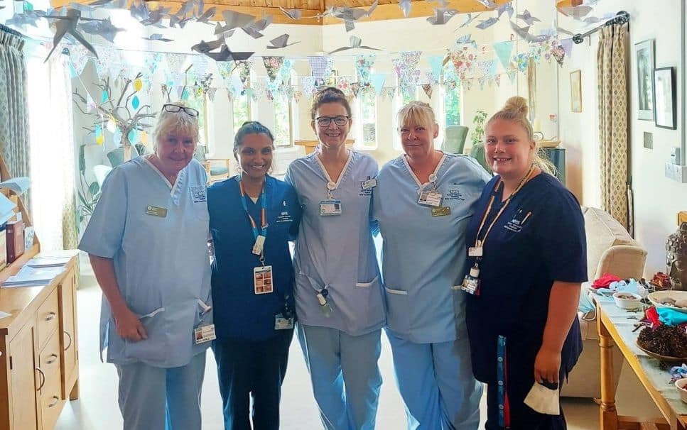Five healthcare workers smiling indoors, wearing uniforms and ID badges.