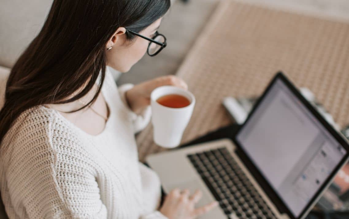 Woman with glasses using a laptop, holding a cup of tea in a cozy setting.