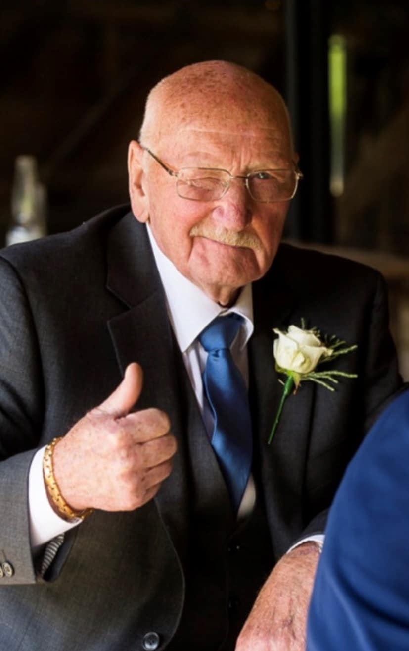Smiling man in a suit giving a thumbs up, wearing glasses and a white rose as a buttonhole.