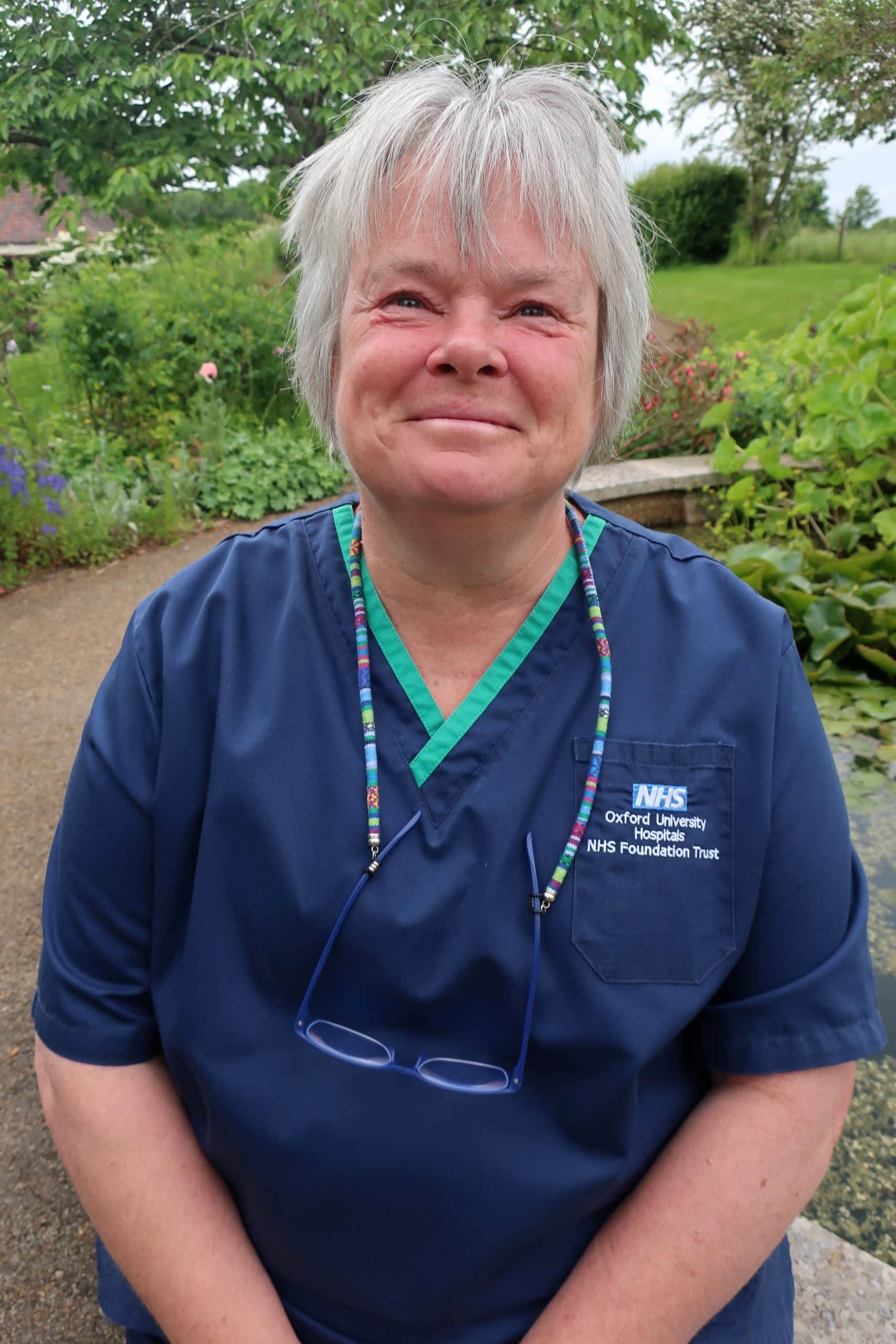 Trish, a palliative care nurse in the Katharine House Hospice blue uniform, smiling and sitting in the hospice gardenwith greenery in the background.