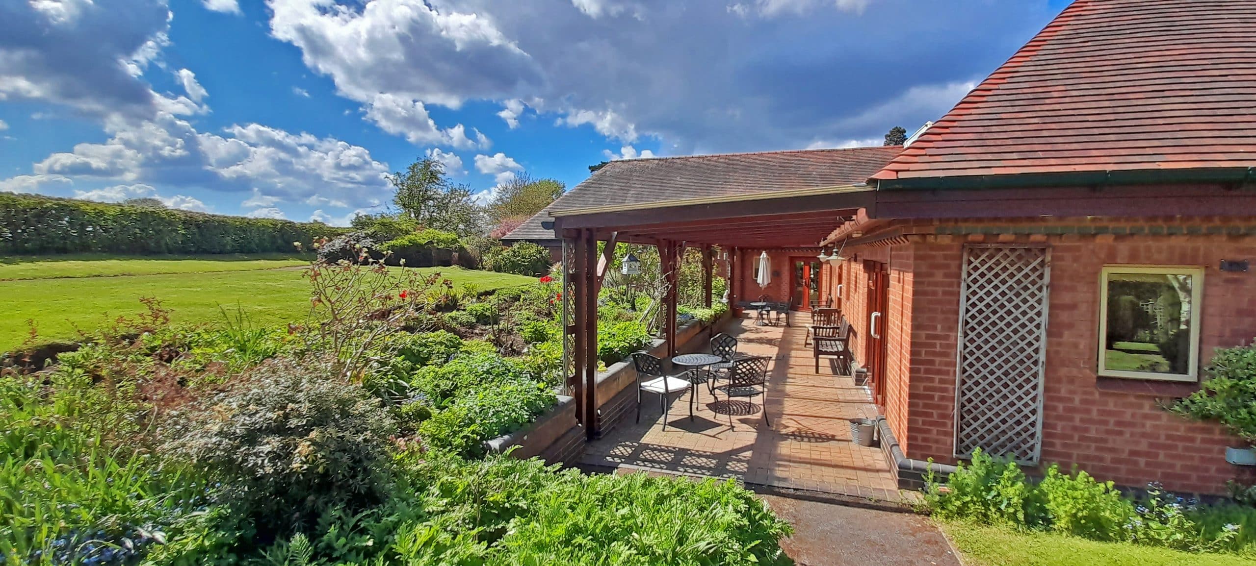 A red-brick building with a covered patio, set beside gardens and open green fields under a blue sky.