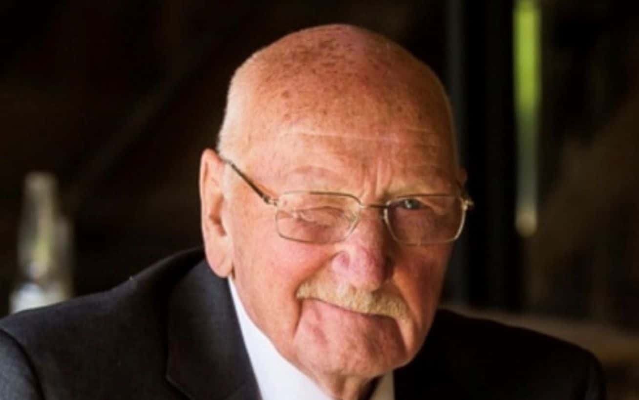 Smiling man with glasses winking, wearing a suit and tie against a dark background.