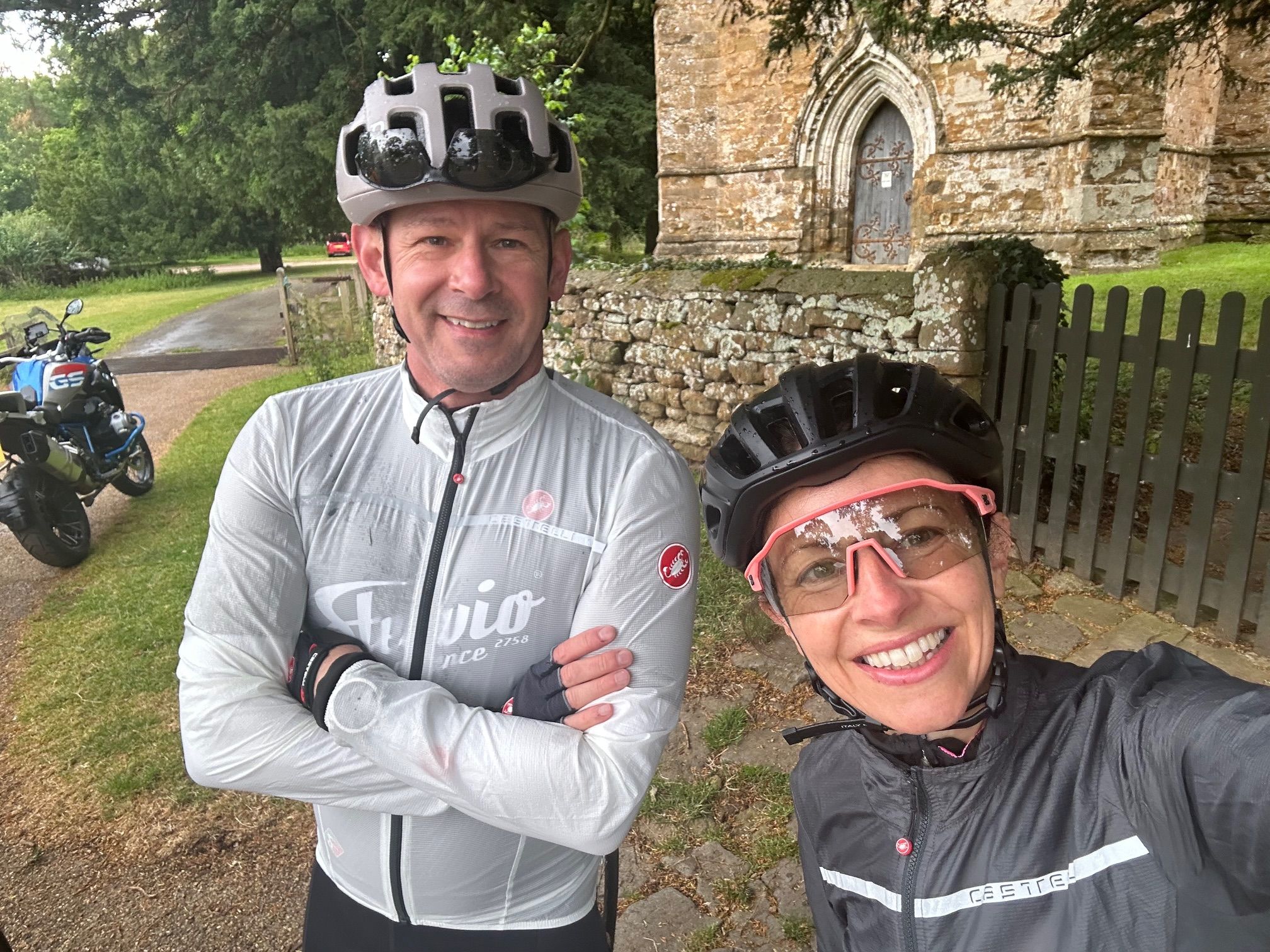 CHRIS&~1 Two cyclists smiling outdoors, wearing helmets and jackets, with a stone building in the background.