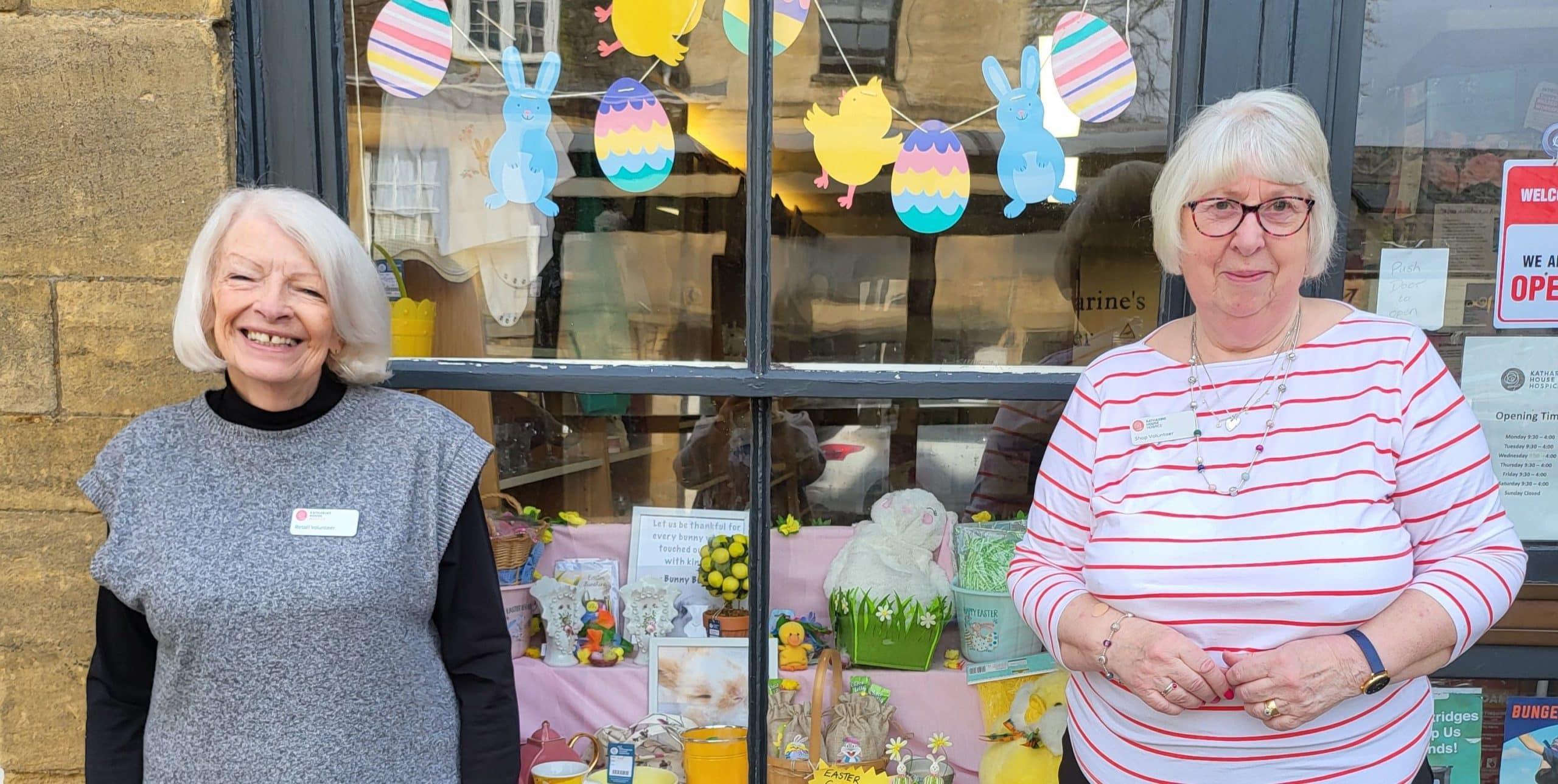 Chipping Norton shop volunteers Two women stand smiling outside a shop with colorful window decorations and cards displayed inside.