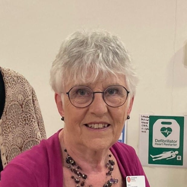 An older person with glasses and short hair, wearing a pink top, stands indoors beside a defibrillator sign.