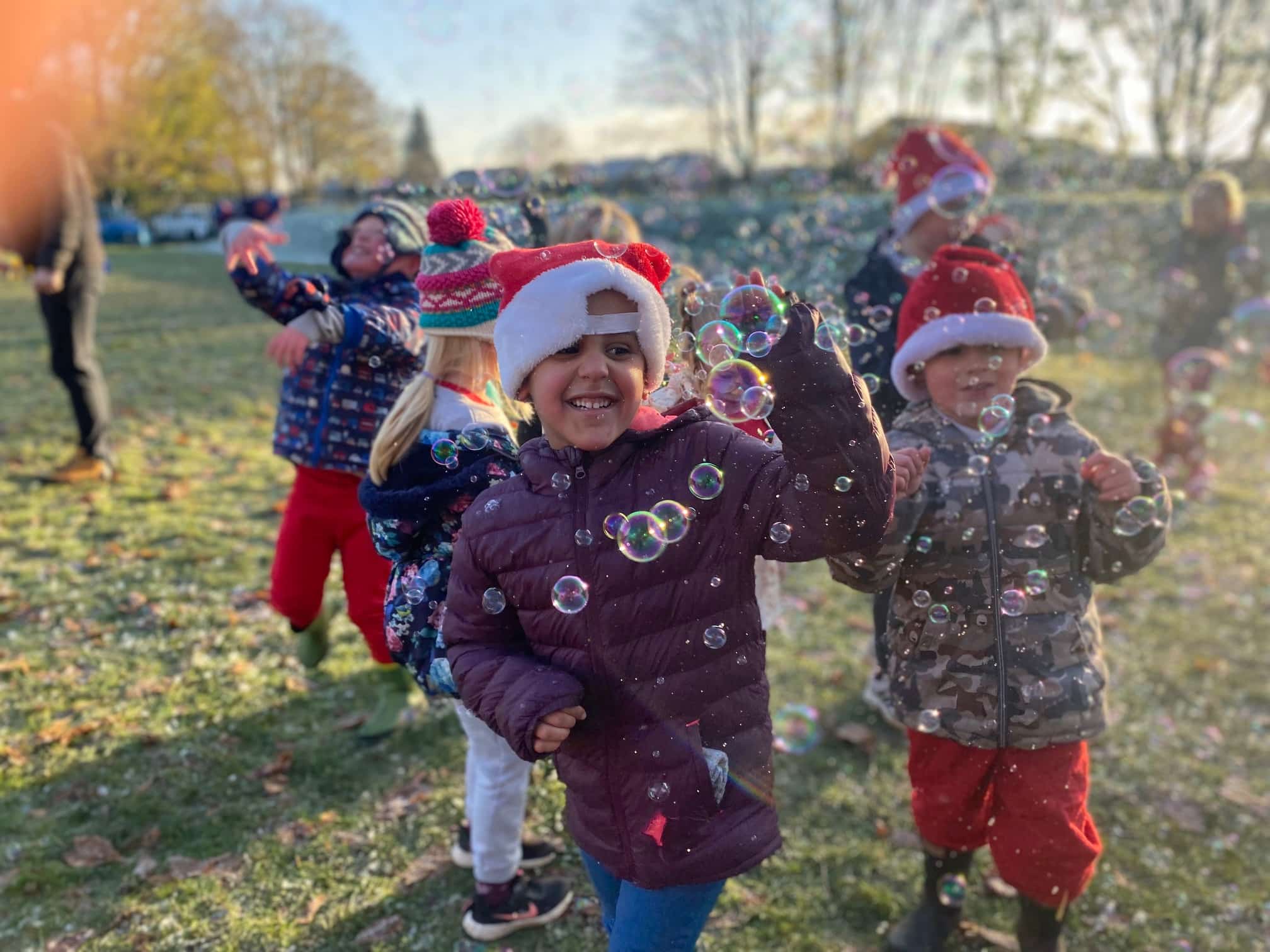 IMG_2329 (2) (1) Children wearing Santa hats playing with bubbles outside on a sunny winter's day after completing the Schools Santa Run challenge for Katharine House Hospice.