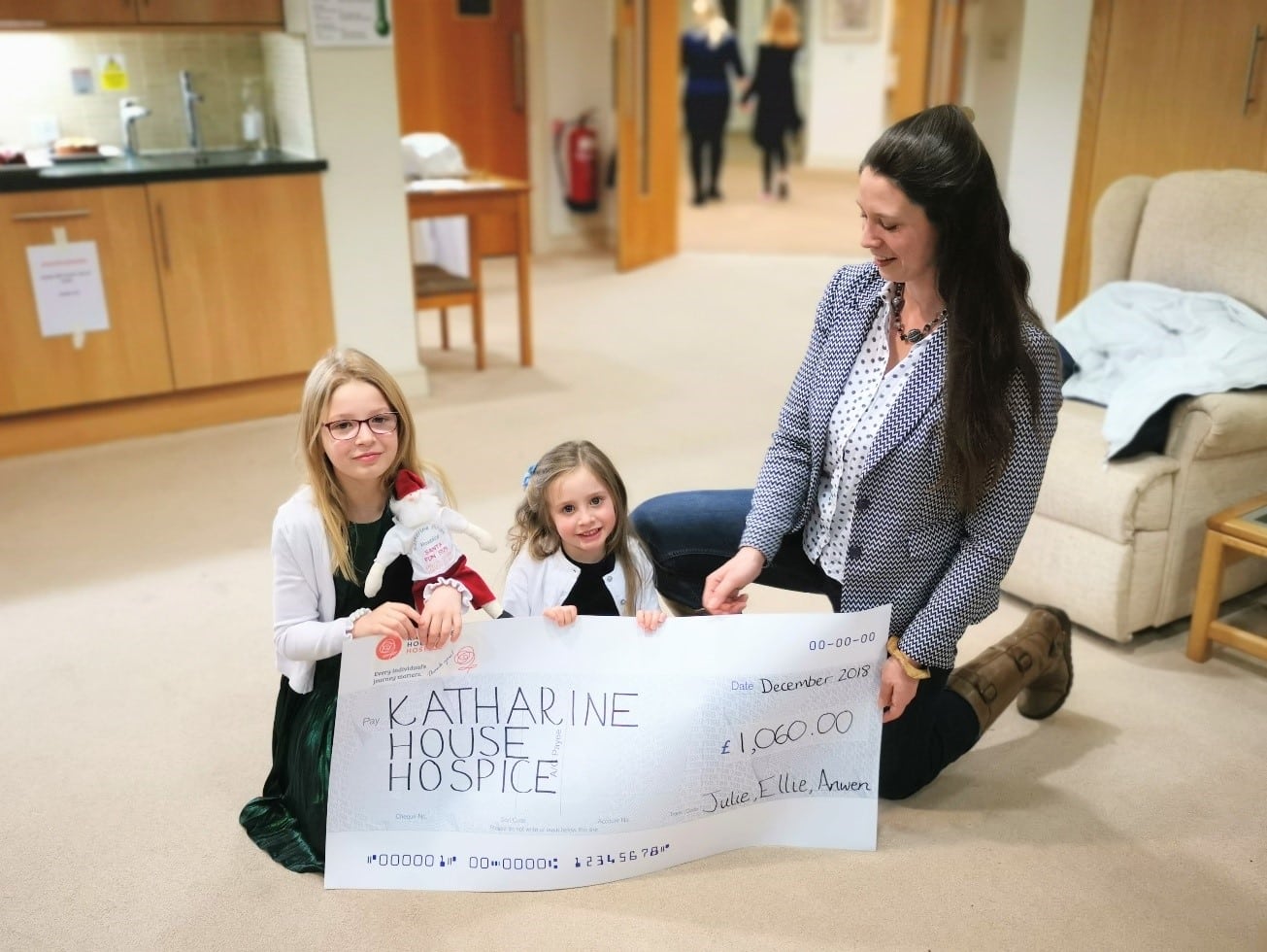 A woman and two children smiling while holding a large donation check to Katharine House Hospice in the Living Well lounge.