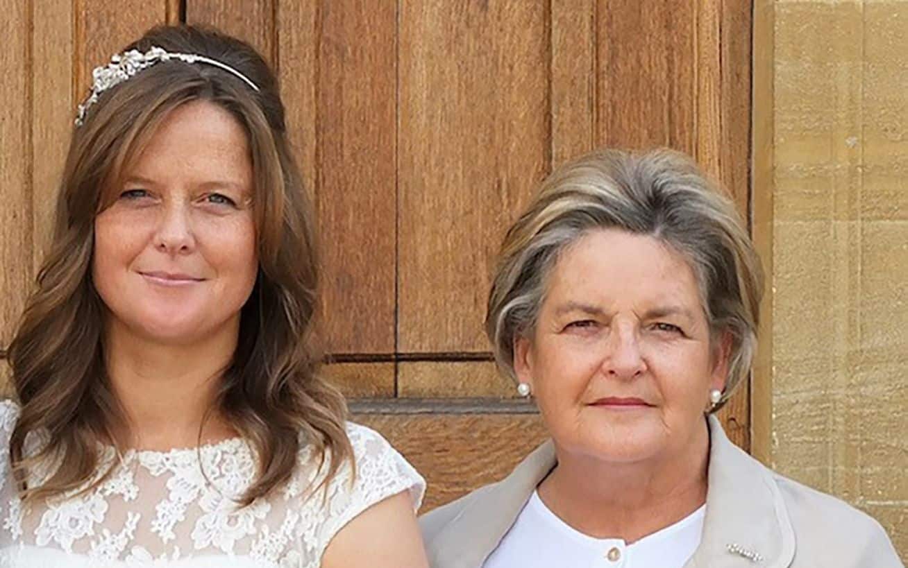 Two women standing in front of a wooden door, one in a lace dress, the other in a beige jacket.