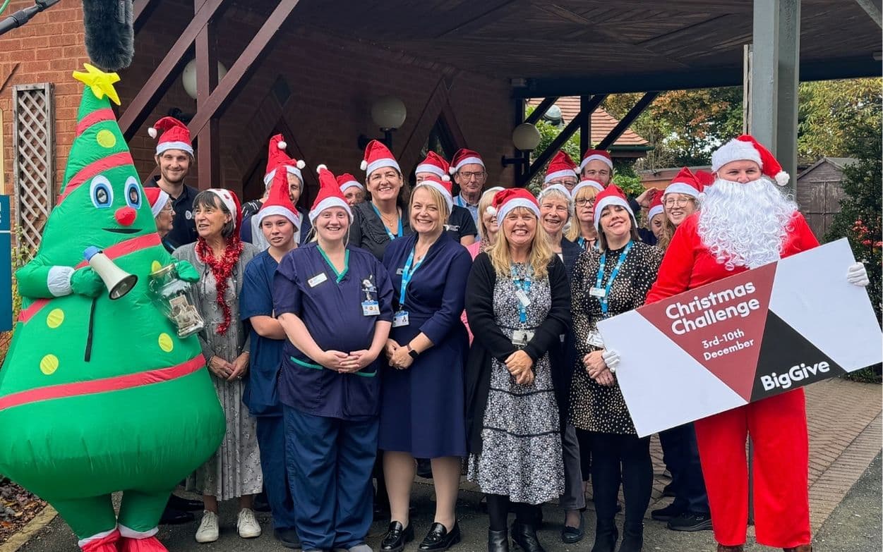 A group of Katharine House Hospice staff stand outside with Father Christmas and a blow-up Christmas Tree with their Christmas hats and a sign reading Christmas Challenge 3rd to 10th December. Big Give.