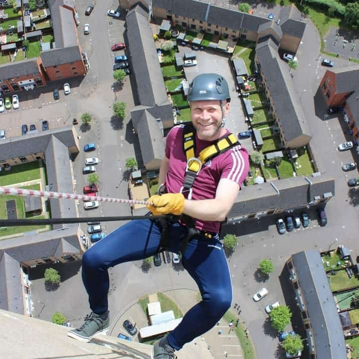 Peter Critchley Person abseiling down a building, smiling, with a view of a housing estate in Northampton below.