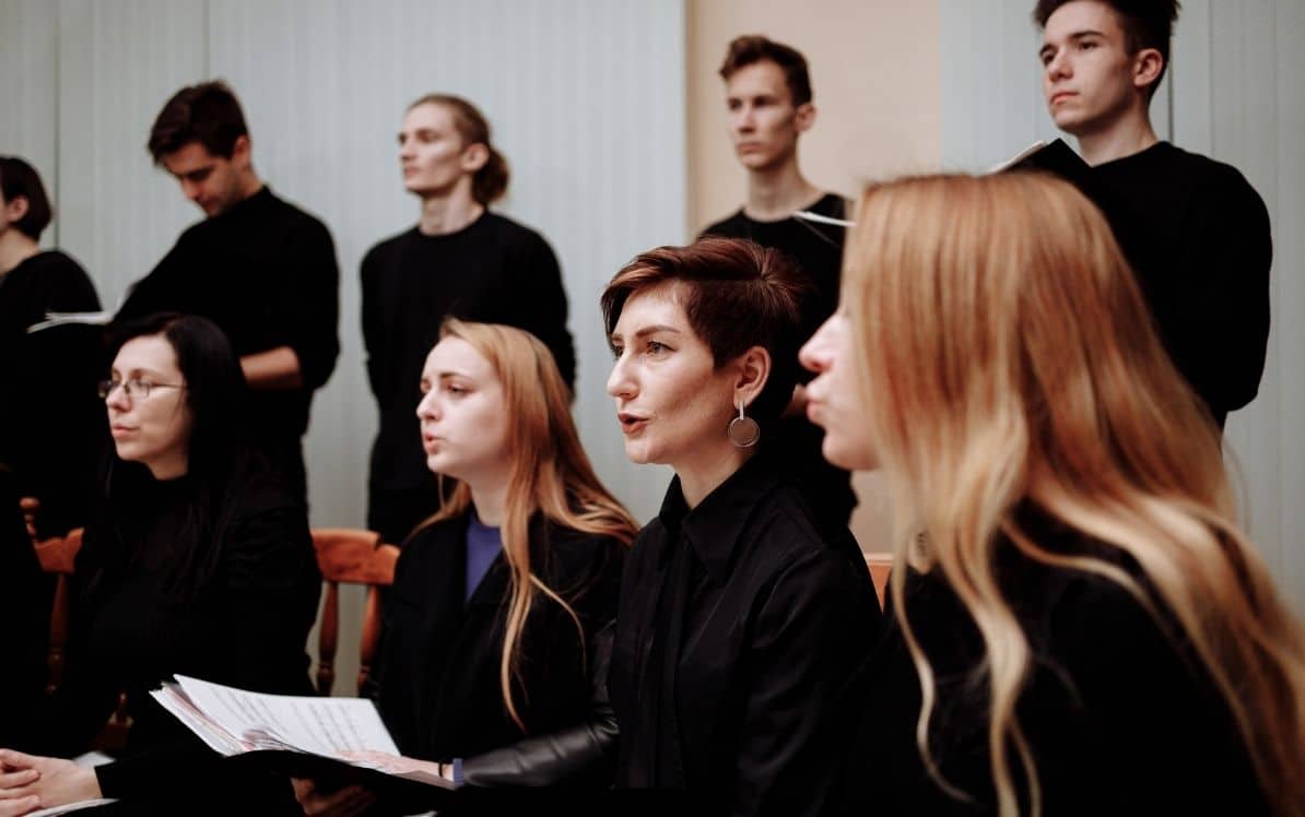 A group of people in black attire singing, some holding sheets of music.