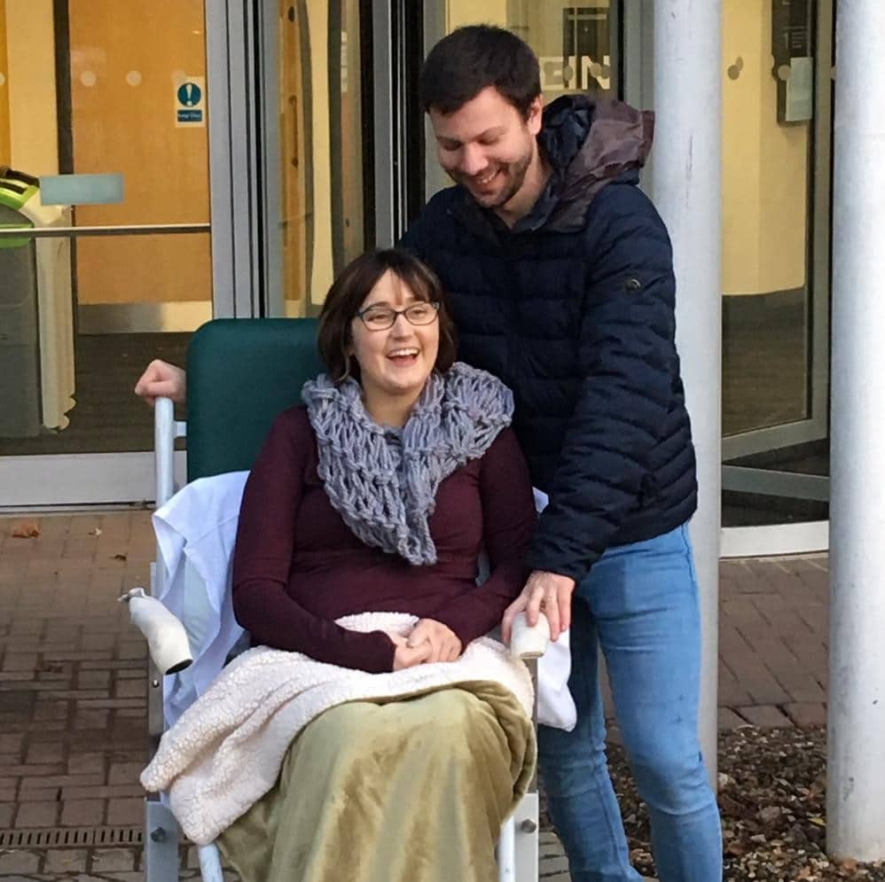 Woman in wheelchair smiling with a man standing behind her outside Katharine House Hospice.
