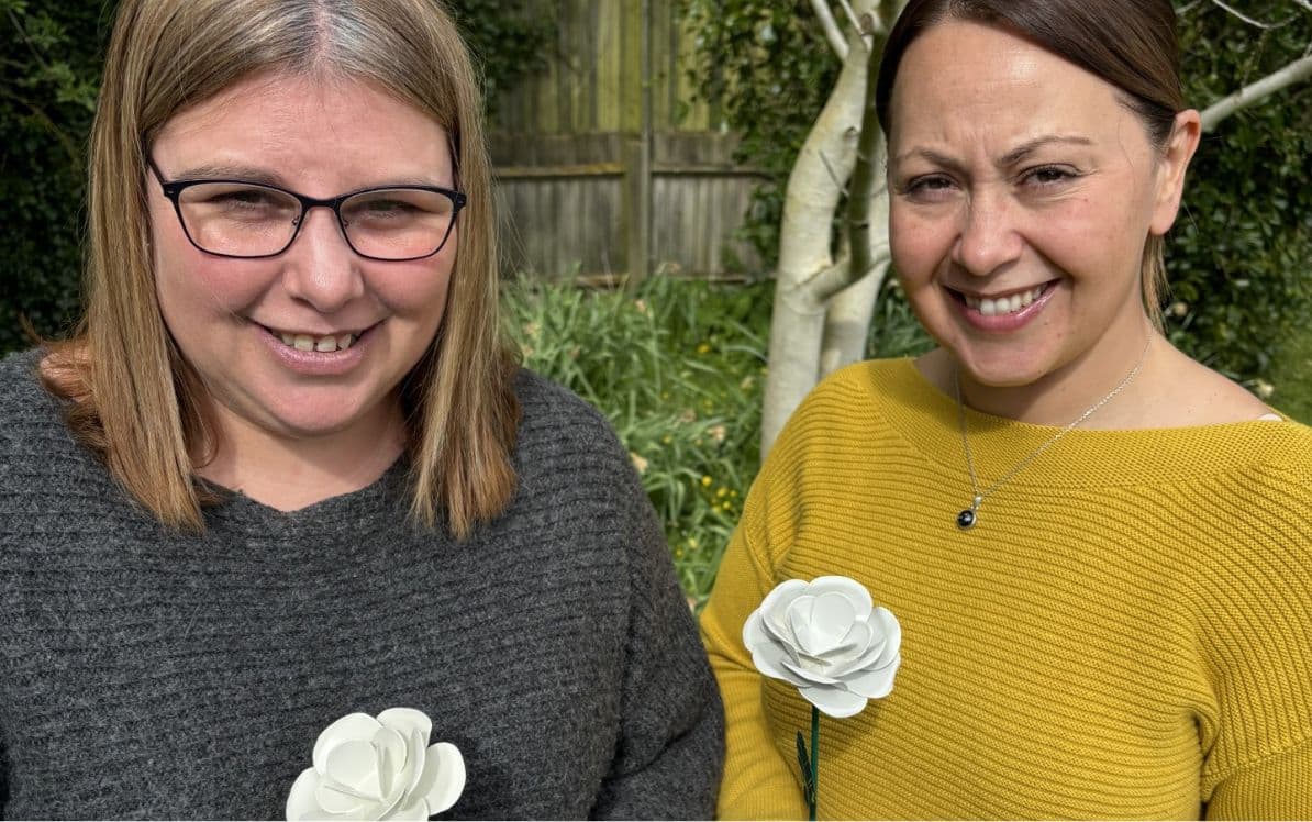 Two women smiling outdoors, each holding a white rose.