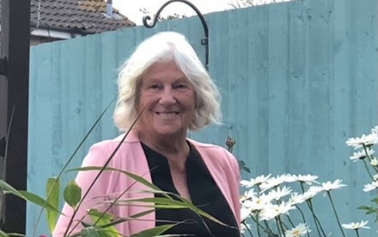Shirley and Linnie Woman smiling in a garden with white flowers and a blue fence in the background.