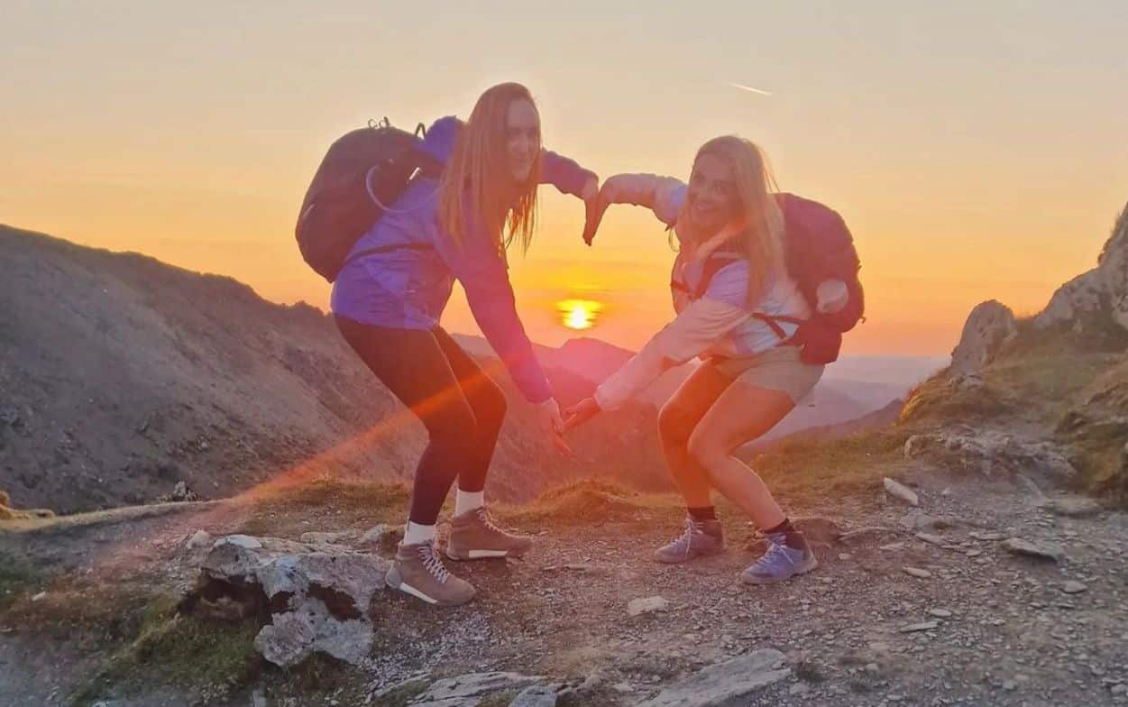 Two hikers at sunrise at the top of Snowdon forming a heart shape with their arms and the sun showing through.