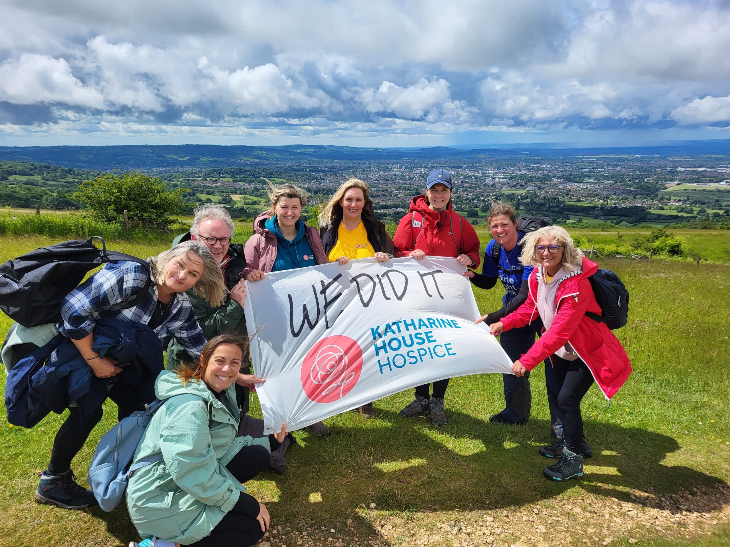 Group holding a "We Did It! Katharine House Hospice" banner on a cloudy and windy day on the top of the Malvern Hills.