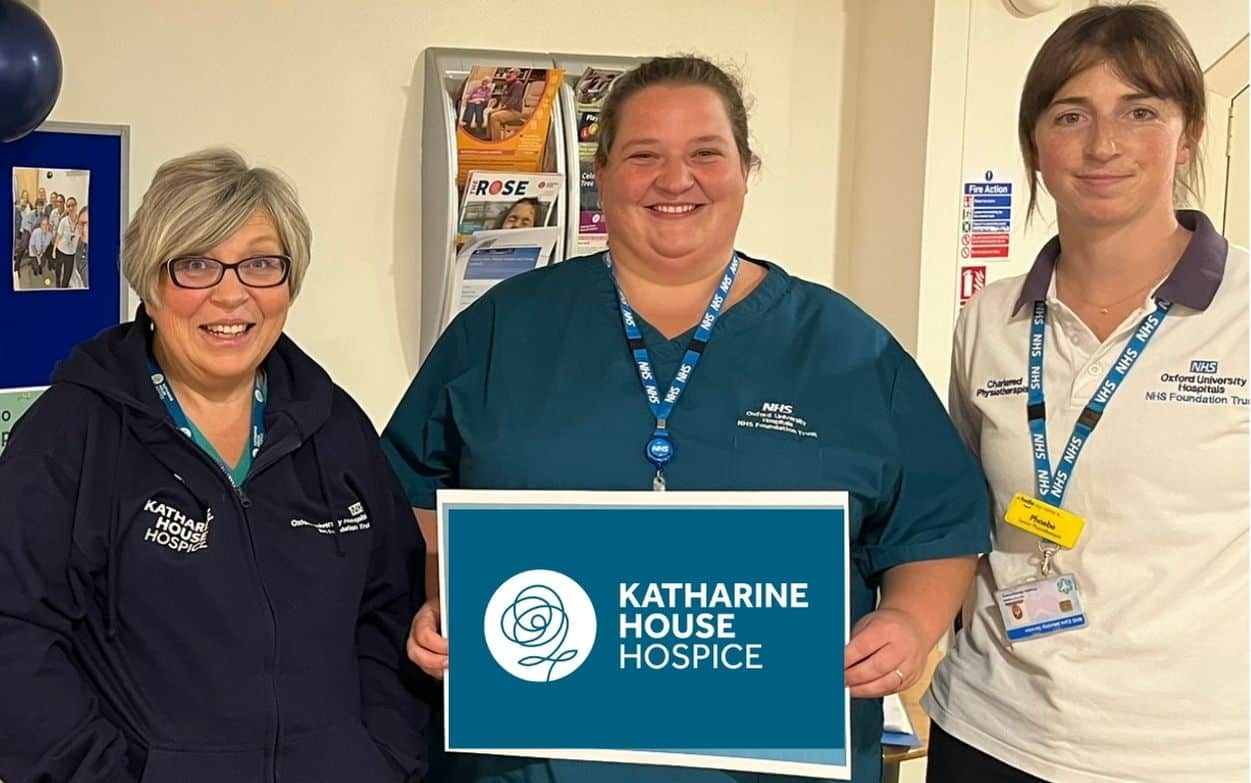 Three smiling women holding a Katharine House Hospice sign indoors.