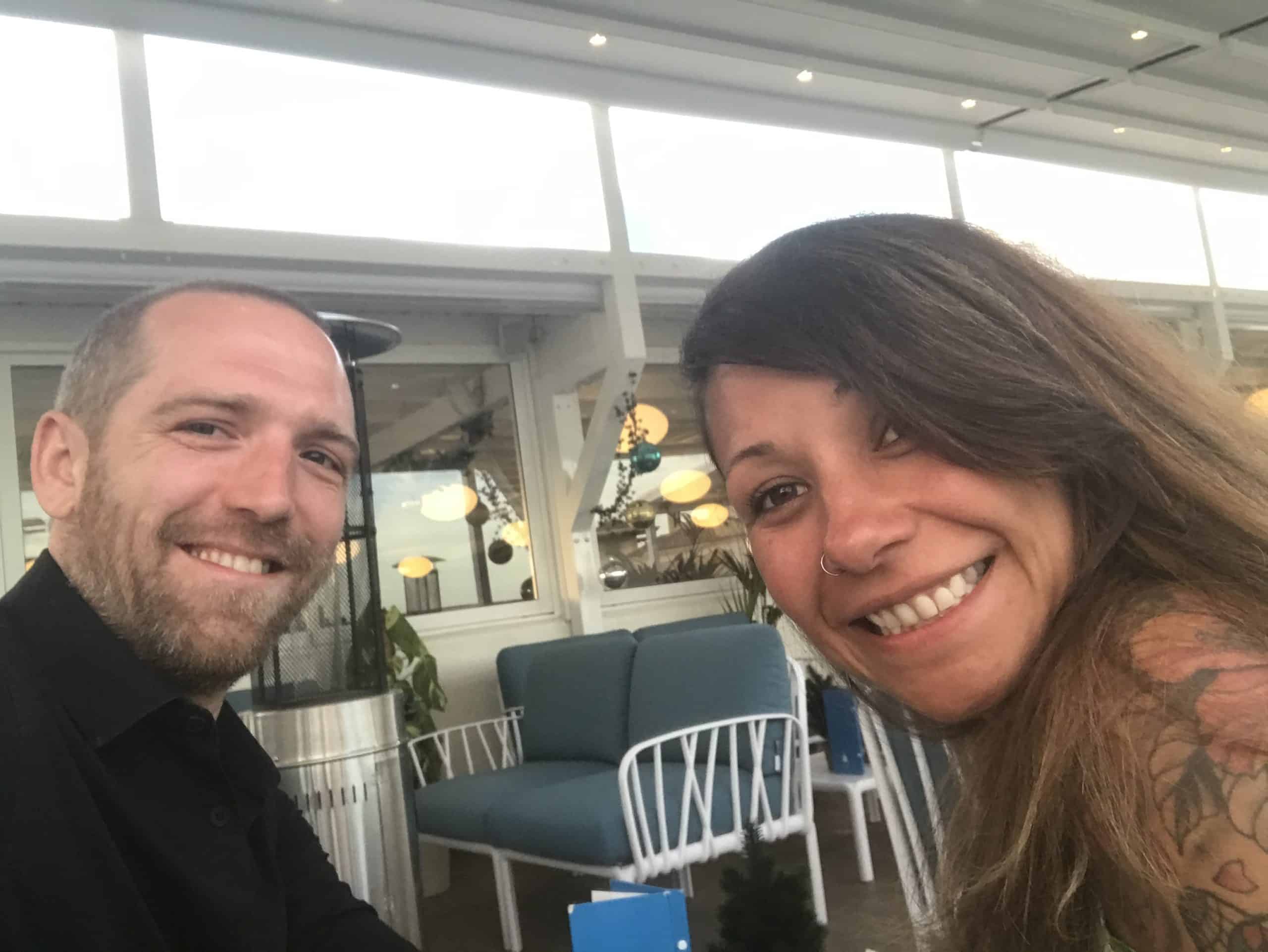 A smiling man and woman sitting in a bright, modern room with blue chairs and decorative lights.