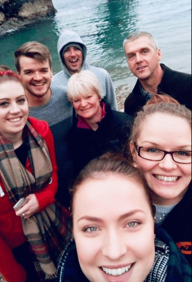 Group selfie by the water, six people smiling outdoors.