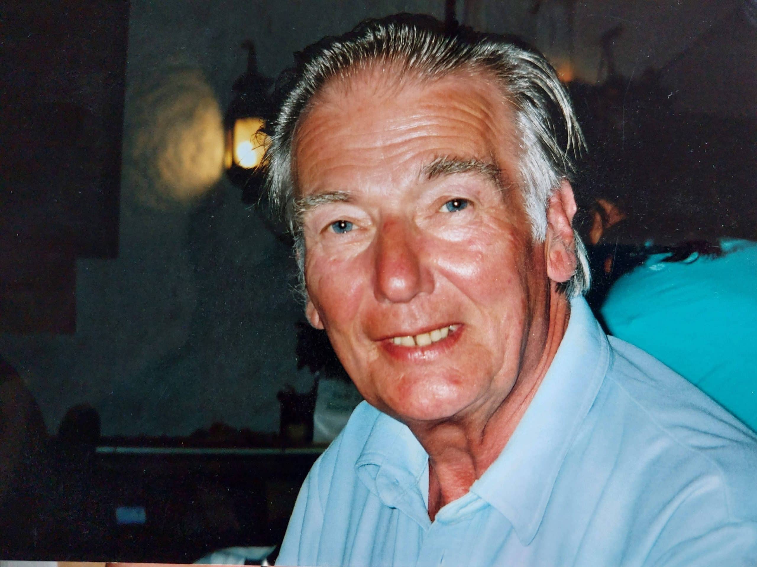 karenturveycasestudy-brian Smiling man with grey hair wearing a light blue shirt indoors.