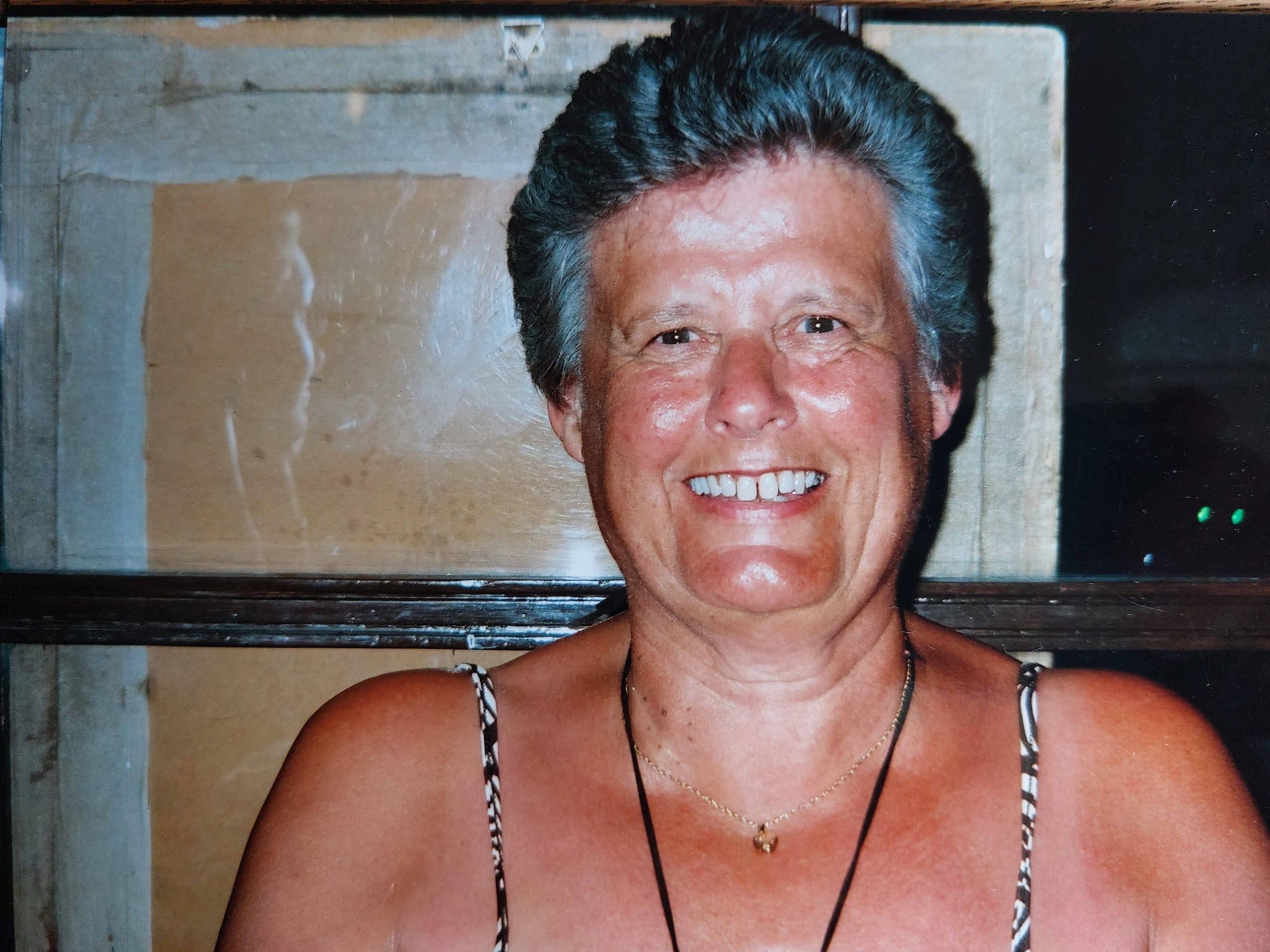 karenturveycasestudy-val Smiling woman with short grey hair, wearing a necklace and sleeveless top, indoors.