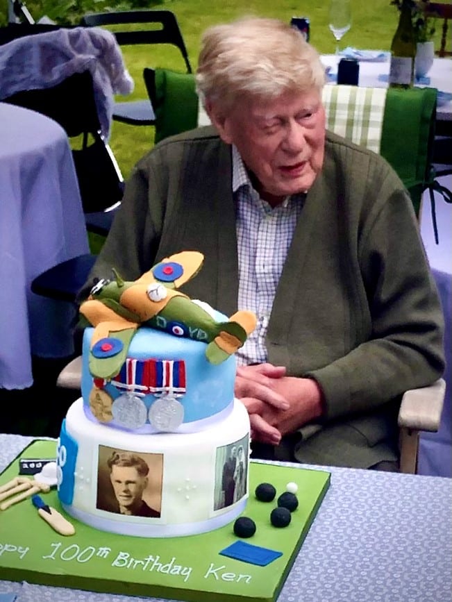 Man sitting outdoors with his 100th birthday cake decorated with a plane and medals.