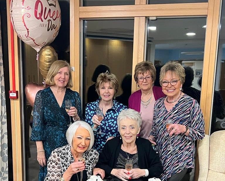 Sue and family Six women smiling and holding drinks, with a "Queen for a Day" balloon in the background.