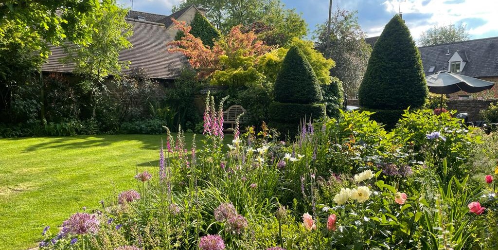 barn house South Newington Open Gardens banner Lush garden with blooming flowers in front of a barn house under a partly cloudy sky.