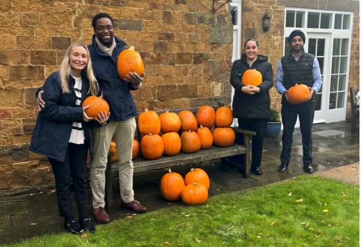 Four people holding pumpkins stand by a bench with more pumpkins outside a building.