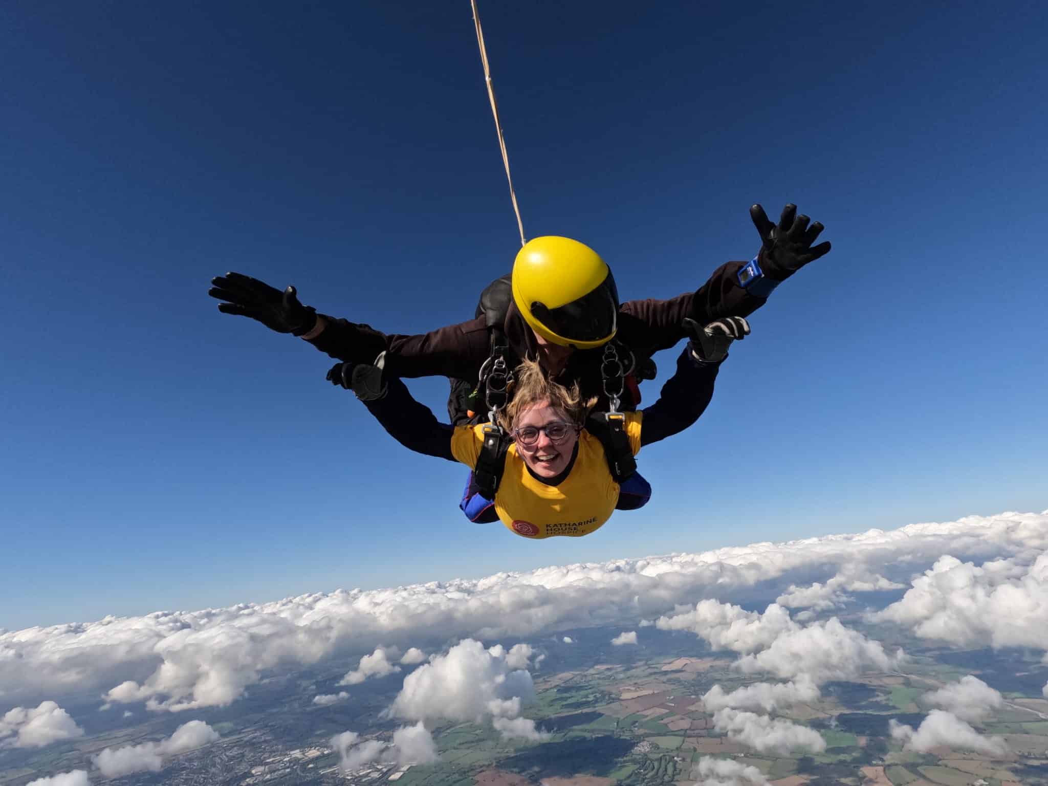 Two people tandem skydiving for Katharine House Hospice above clouds on a clear, sunny day.