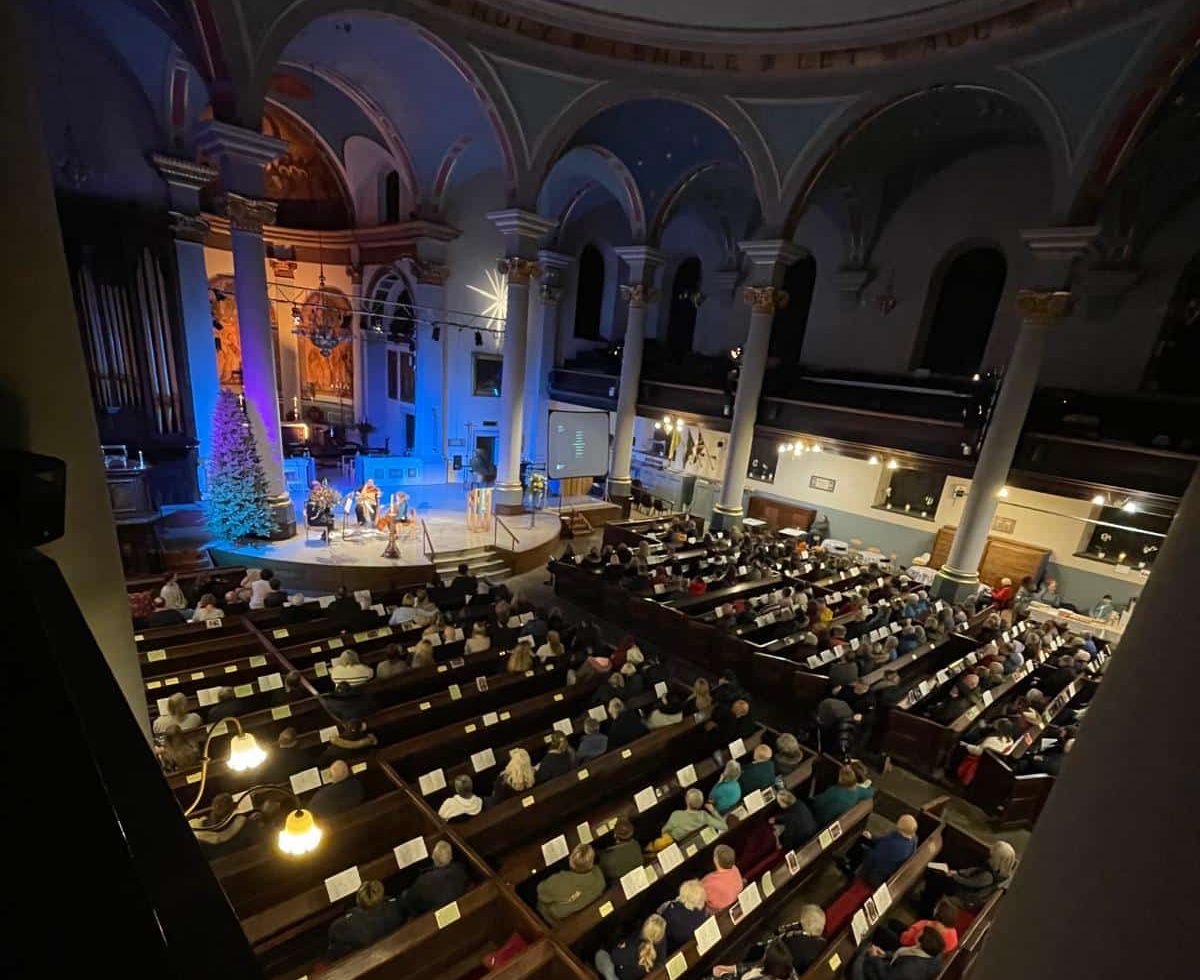 An audience sits inside St Mary's church in Banbury during the Lights of Love evening watching a stage lit with blue and orange lights.