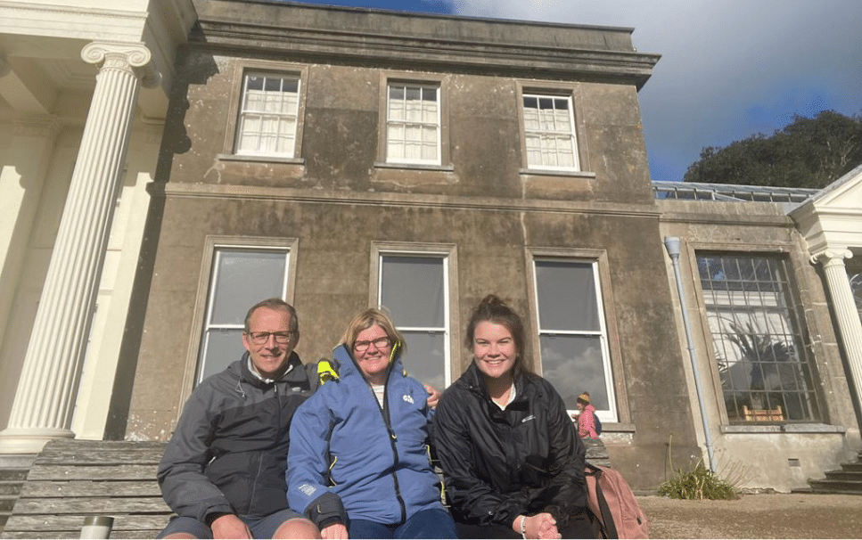 A man with his arm around one woman and a second woman sitting alongside in front of a stately home smiling at the camera.