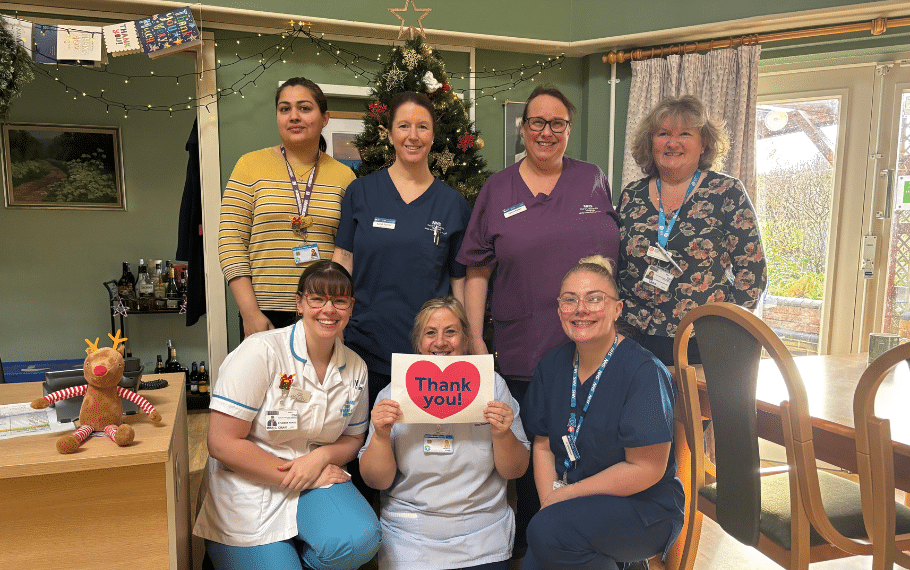 A group of nurses from Katharine House Hospice stand in two rows, smiling and holding a sign with a heart and 'Thank you' written in its centre.