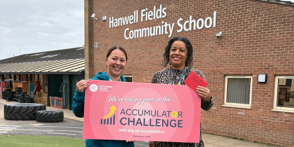 Schools Challenges And Events Two women stand in front of Hanwell Fields Community School holding a sign that says 'We're taking part in the Accumulator Challenge'.