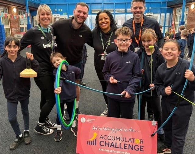 Hanwell-Fields-Community-School-students-and-staff-1 A group of primary school children and staff grouped together holding sports equipment and a sign that says 'We're taking part in the Accumulator Challenge'.